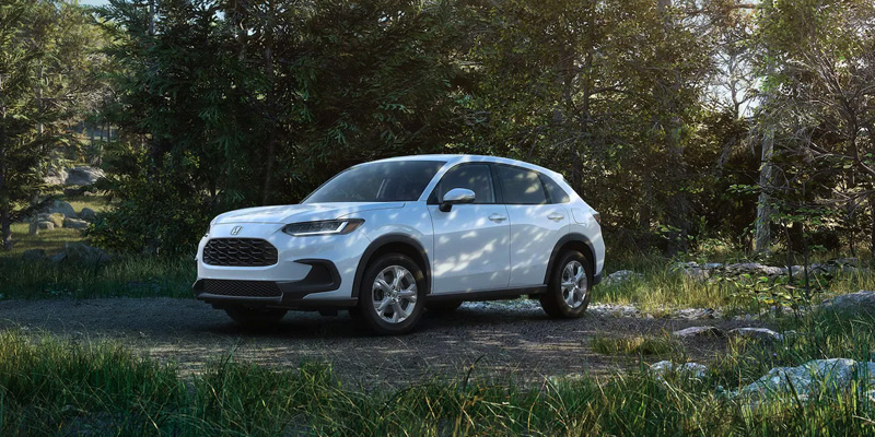 Front three-quarter view of a white compact SUV parked on a dirt trail in a lush forest setting with sunlight filtering through the trees.