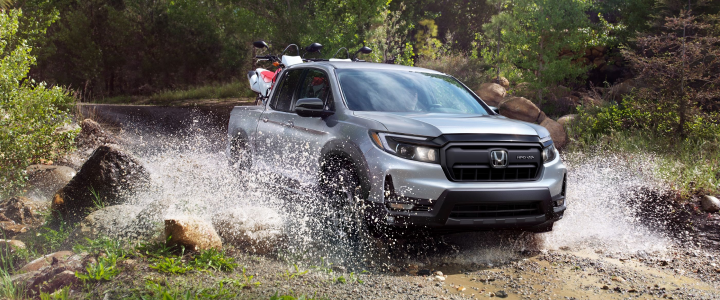 Silver Honda Ridgeline splashing through a rocky creek crossing with dirt bikes in its bed