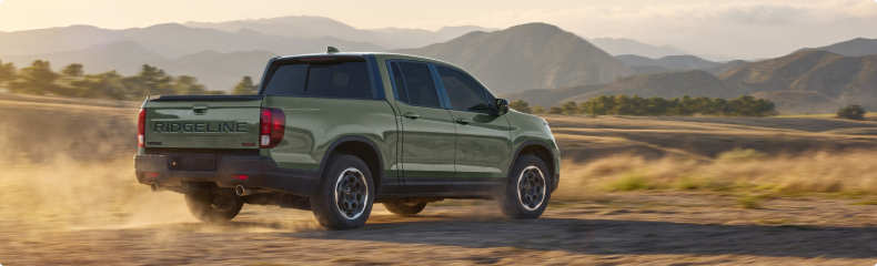 Green Honda Ridgeline TrailSport kicking up dust on a dirt road below mountains, seen from the rear quarter
