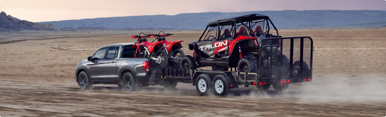 Grey Honda Ridgeline towing a trailer loaded with a Honda Talon side-by-side across a desert, with dirt bikes in its bed
